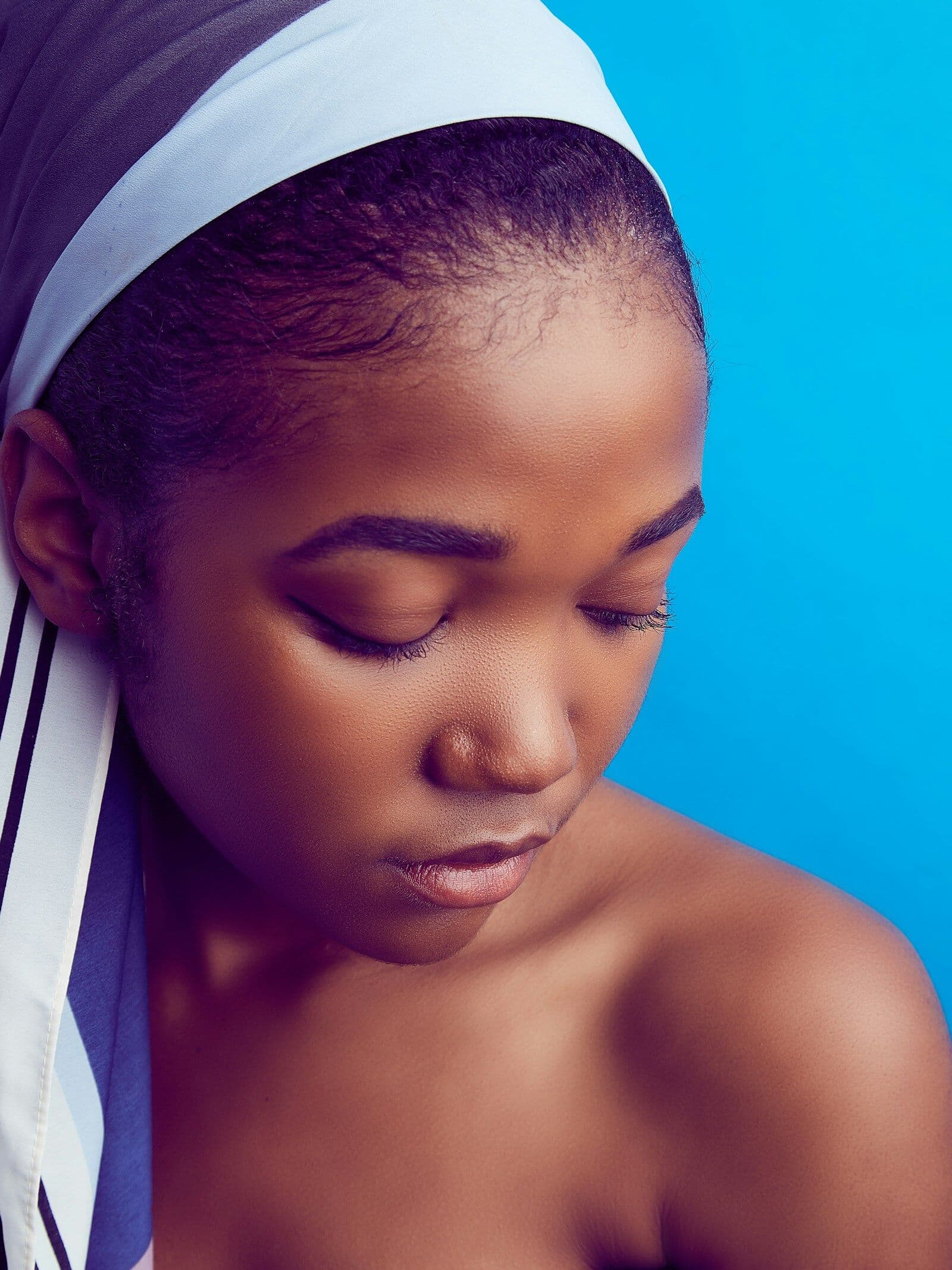 Woman in a white headscarf looking down softly against a bright blue backdrop
