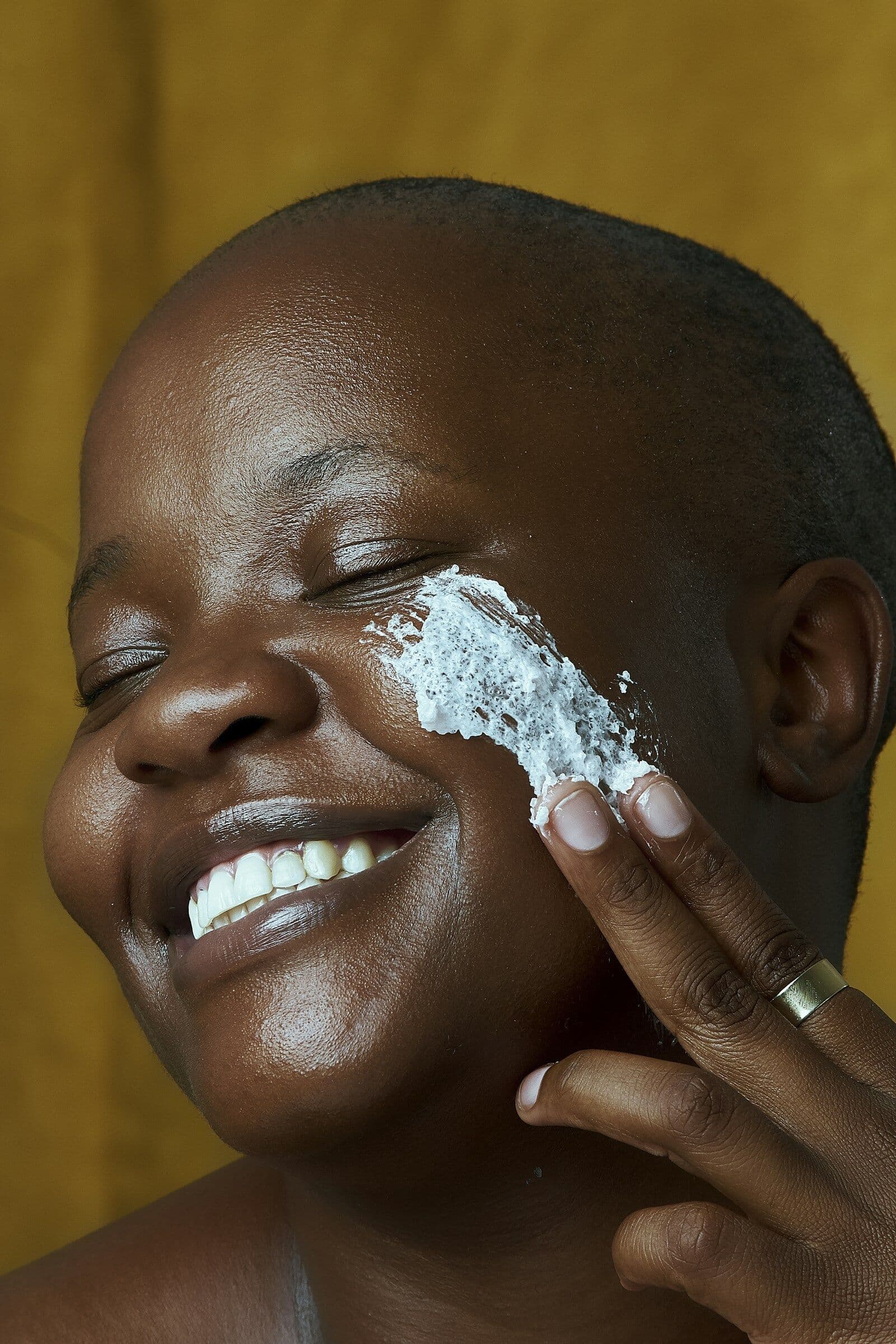 Laughing woman with a shaved head applying a cleansing mask to her cheek