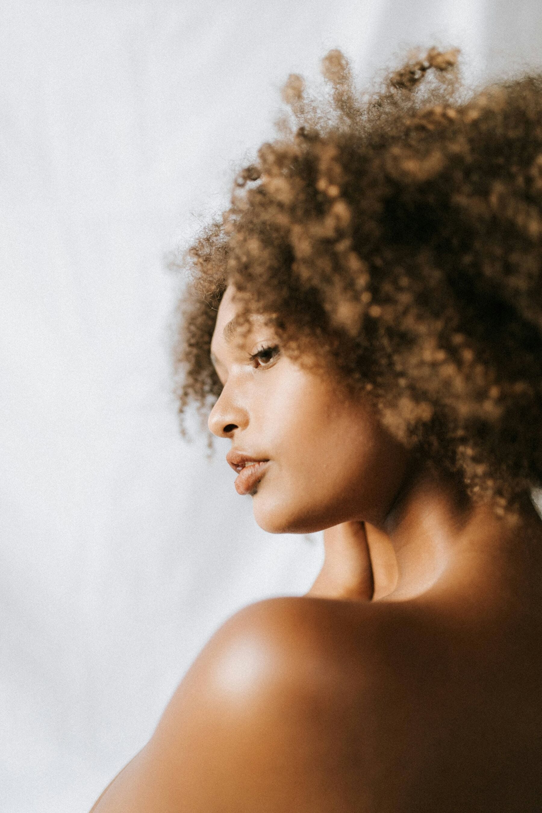 Black woman with natural curly hair and softly glowing skin, photographed in profile against a bright studio backdrop