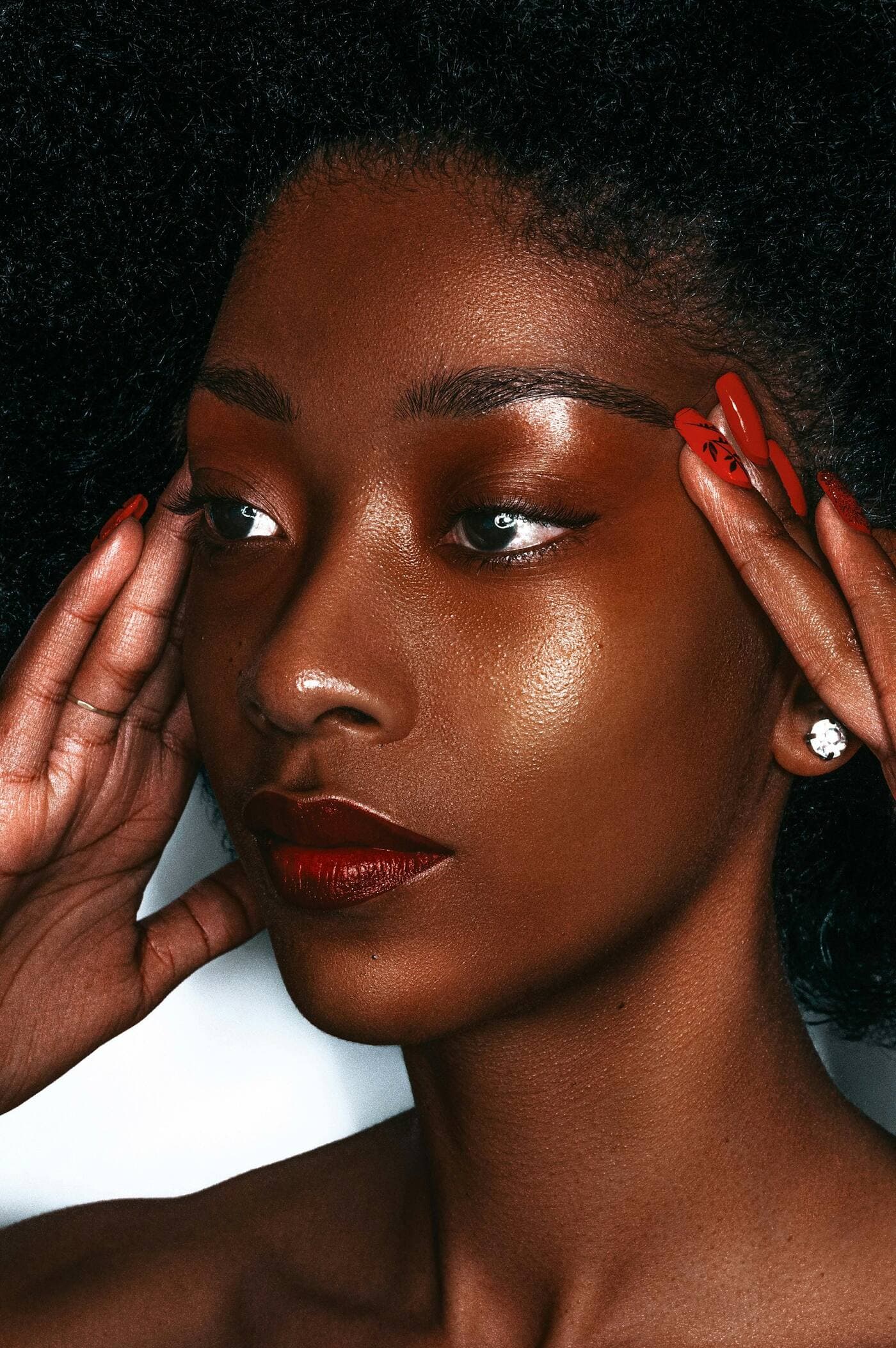 Woman with deep brown, glowing skin resting her fingertips at her temples, showing a glossy red manicure